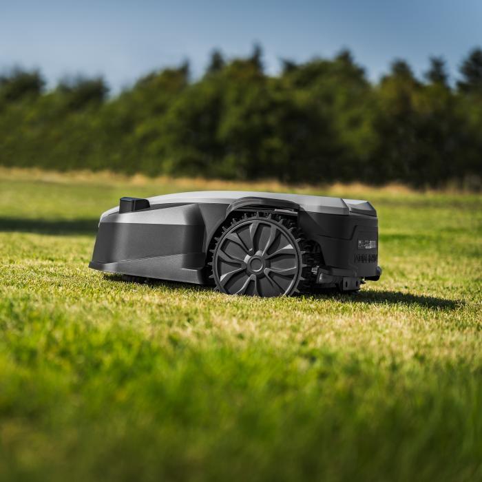 Grå og sort robotplæneklipper kører på en stor græsplæne med træer i baggrunden